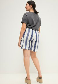 Šedé tričko s krátkým rukávem, které je kombinováno se šortkami s modro-bílým pruhovaným vzorem, vybavenými kapsami a uvolněným střihem. Na nohou má béžové sandály na platformě.