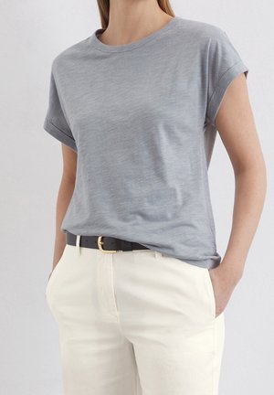 Frau trägt ein hellgraues kurzärmeliges T-Shirt, in weiße Hose mit schwarzem Gürtel gesteckt, Hände in den Taschen, neutraler Hintergrund.