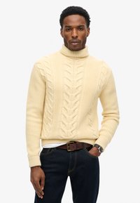 Crème kleurige kabelgebreide coltrui met ribgebreide zomen, gecombineerd met donkere jeans en een bruine riem. Heeft een gestructureerd, warm ontwerp.