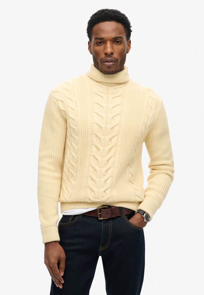 Cremfarbener Rollkragensweater mit Kabelstrickmuster und gerippten Abschlüssen, kombiniert mit dunklen Jeans und einem braunen Gürtel. Verfügt über ein strukturiertes, warmes Design.
