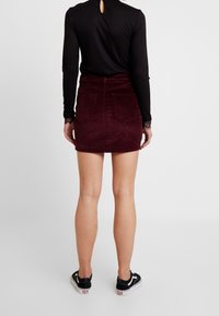 Jupe en velours côtelé bordeaux avec deux poches arrière, taille haute et une encolure au-dessus de mi-cuisse. Associée à un haut à manches longues noir et des baskets.