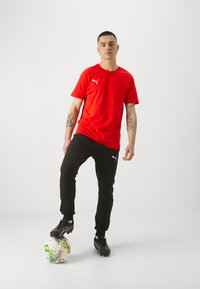 Rode sportieve t-shirt, zwarte joggerbroek met opgerolde enkels en zwarte voetbalschoenen. Toont een kleurrijk patroon voetbal aan de voet.