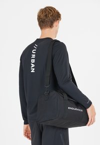 Musta duffel-laukku säädettävällä olkahihnalla, jossa on pyöreä muoto, kestävä kangas ja valkoinen teksti "ENDURANCE" sivussa.
