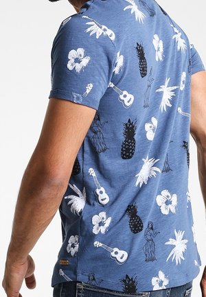 Uomo che indossa una maglietta blu con stampe tropicali bianche e nere di ananas, fiori di ibisco, chitarre, palme e ballerini di hula.