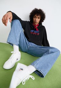 Felpa nera con testo colorato, jeans blu, sneakers bianche e un bandana rosso. La texture è casual e rilassata, posta su una superficie verde.