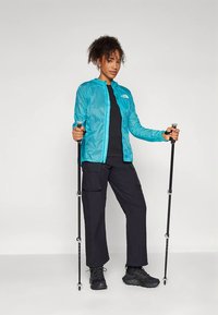 Kvinde i en turkise jakke og sorte cargo-bukser, der holder trekkingstave, står klar til vandretur eller udendørs aktivitet.
