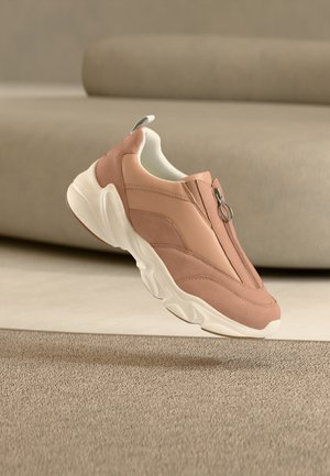 Baskets rose clair avec semelle blanche épaisse et fermeture éclair à l'avant, flottant au-dessus d'un tapis beige et d'un canapé beige assorti en arrière-plan.