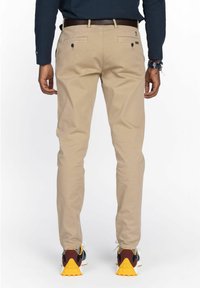 Mann in beigen, schmal geschnittenen Hosen, schwarzem Gürtel, navy-blauem Langarmhemd, Armbändern und mehrfarbigen Sneakers mit gelben Sohlen, der mit dem Rücken zur Kamera steht.