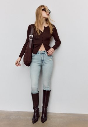 Kobieta w ciemnobrązowym kardiganie na guziki, jasnoniebieskich jeansach z wysokim stanem i przetarciami, ciemnobrązowych kozakach za kolano oraz okularach przeciwsłonecznych, stojąca w pomieszczeniu.