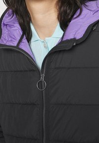 Zwarte pufferjas met een paarse voering, voorzien van een ritssluiting en een ronde trekker. Daaronder is een lichtblauw overhemd met kraag zichtbaar.