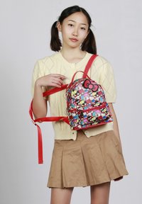 Mochila roja con patrones coloridos de Hello Kitty, textura suave, forma redondeada y un prominente detalle de lazo negro en la parte superior.