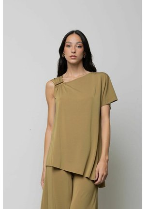 Donna con lunghi capelli scuri che indossa orecchini a cerchio dorati, una maglia beige asimmetrica con una manica corta e pantaloni beige abbinati.