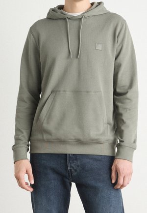 Sudadera de algodón gris con cordón, bolsillo frontal y parche con logo. Textura suave, puños y dobladillo acanalados. Combinada con jeans azul oscuro.