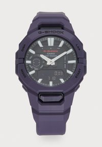 Montre G-Shock violette avec un cadran noir rond, des index horaires blancs et un affichage numérique. Elle est équipée d'un bracelet en caoutchouc et de plusieurs boutons de fonction.