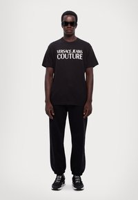 Melnas īsām piedurknēm T-kontē ar "VERSACE JEANS COUTURE" logotipu, apvienota ar melnām sporta biksēm un melniem sporta apaviem ar baltajiem akcentiem.