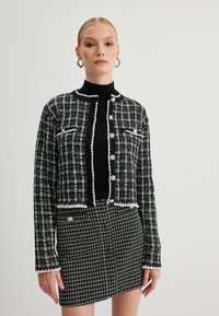 Musta-valkoinen ruudullinen tweed-takki, jossa on lyhyt malli, hopeanappiset yksityiskohdat ja rispaantunut reuna. Yhdistetty vastaavaan hameeseen.