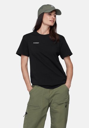 Femme portant un t-shirt noir Mammut, un pantalon vert olive avec des poches, et une casquette de baseball vert olive, debout les mains dans les poches.