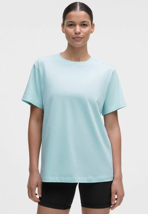 Młoda kobieta z włosami związanymi do tyłu, ubrana w jasnoniebieski krótki rękaw i czarne spodenki kolarskie, stojąca na jednolitym tle.