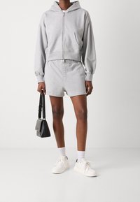 Sudadera gris con cremallera frontal y capucha, combinada con pantalones cortos a juego. El modelo sostiene un pequeño bolso negro. El conjunto incluye zapatillas blancas.