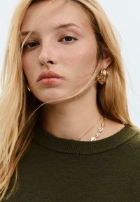 Blondynka z jasnymi piegami nosi złote kolczyki w kształcie kół, złoty naszyjnik i oliwkową sweter z dzianiny, z włosami częściowo zasłaniającymi jej twarz.