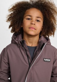 Braune Kapuzenjacke aus glattem Material mit kontrastierendem Reißverschluss und schwarchem Logopatch. Verfügt über einen Reißverschluss vorne und einen verstellbaren Kragen.