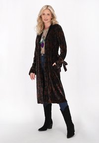 Lungo kimono di velluto scuro con motivo paisley, legato in vita, con maniche a campana. Indossato sopra una maglietta chiara e jeans blu, con stivali neri.