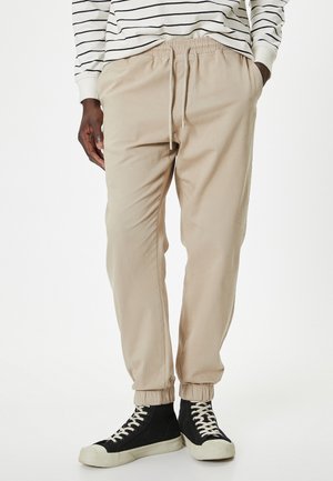 Beige joggerbukser lavet af blødt stof, med elastisk talje og snøre, ribbede ankler og sidelommer.