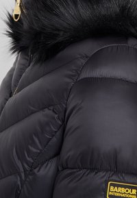 Schwarze Steppjacke mit einer Kapuze mit Pelzbesatz, gestepptem Muster, goldenen Beschlägen und einem Markenpatch an der unteren Seite.