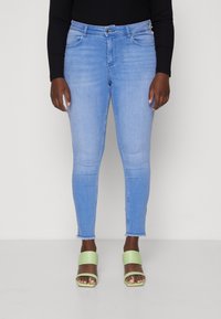 Ljust blå skinny jeans med fransad nederkant, tillverkade av denimtyg. Matchat med gröna blockklack-sandaler.