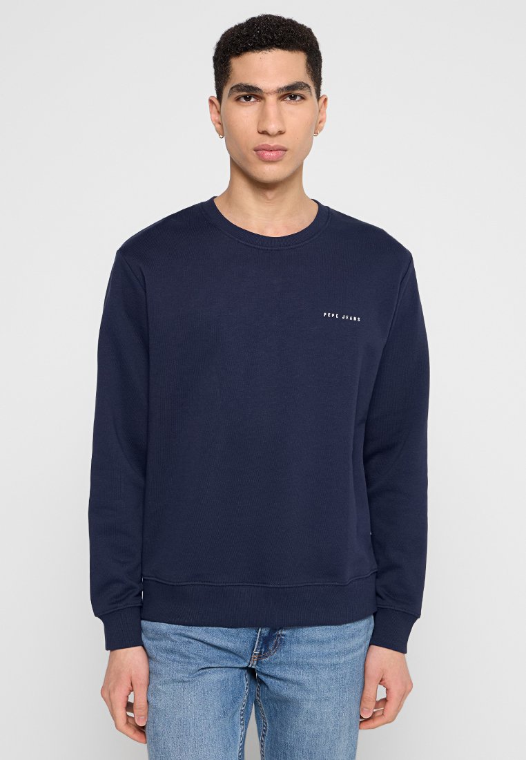 Dunkelblaues Crewneck-Sweatshirt aus geripptem Material mit einem kleinen weißen Logo auf der Brust. Lange Ärmel und gerader Saum. Darunter werden Jeans aus Denim getragen.