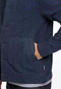 Marineblauwe jas met een soepele textuur, voorzien van een borstzak, zijzakken en een knoopdetail aan de onderkant. Lange mouwen zichtbaar.