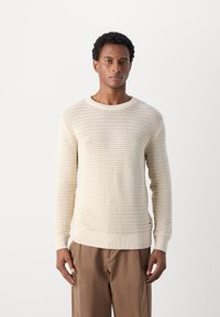 Pull en tricot beige clair avec un motif de tricot ajouré, col rond et poignets et ourlet à côtes. Porté avec un pantalon marron.
