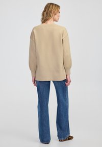 Beżowy, oversize sweter z długimi, lekko bufkowymi rękawami, w połączeniu z niebieskimi dżinsami w stylu dzwonów. Sweter ma prążkowany dekolt i wykończenie.