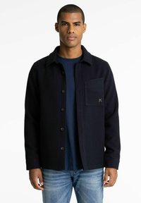 Marineblauwe knoopjacket met een gestructureerde stof, voorzak en zwarte knopen, gelaagd over een donker shirt, gedragen met lichtblauwe spijkerbroek.