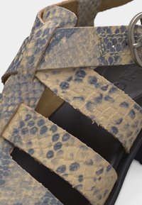 Λουριά σανδαλιού με υφή δέρματος φιδιού σε μπεζ και γκρι αποχρώσεις, με ασημί πόρπη και μαύρη σόλα, κοντινή λεπτομέρεια των αλληλοεπικαλυπτόμενων λουριών και των ραφών.