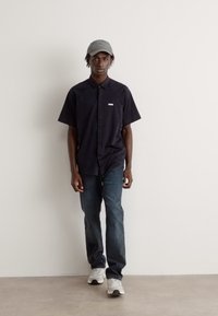 Camisa preta de botão de mangas curtas com detalhe de logo, combinada com jeans escuros e um boné cinza, complementada por ténis brancos. Design simples.