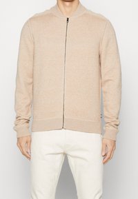 Maglione beige con zip, colletto e polsini a costine, realizzato in un tessuto a maglia testurizzata e dotato di una cerniera metallica visibile sul davanti.