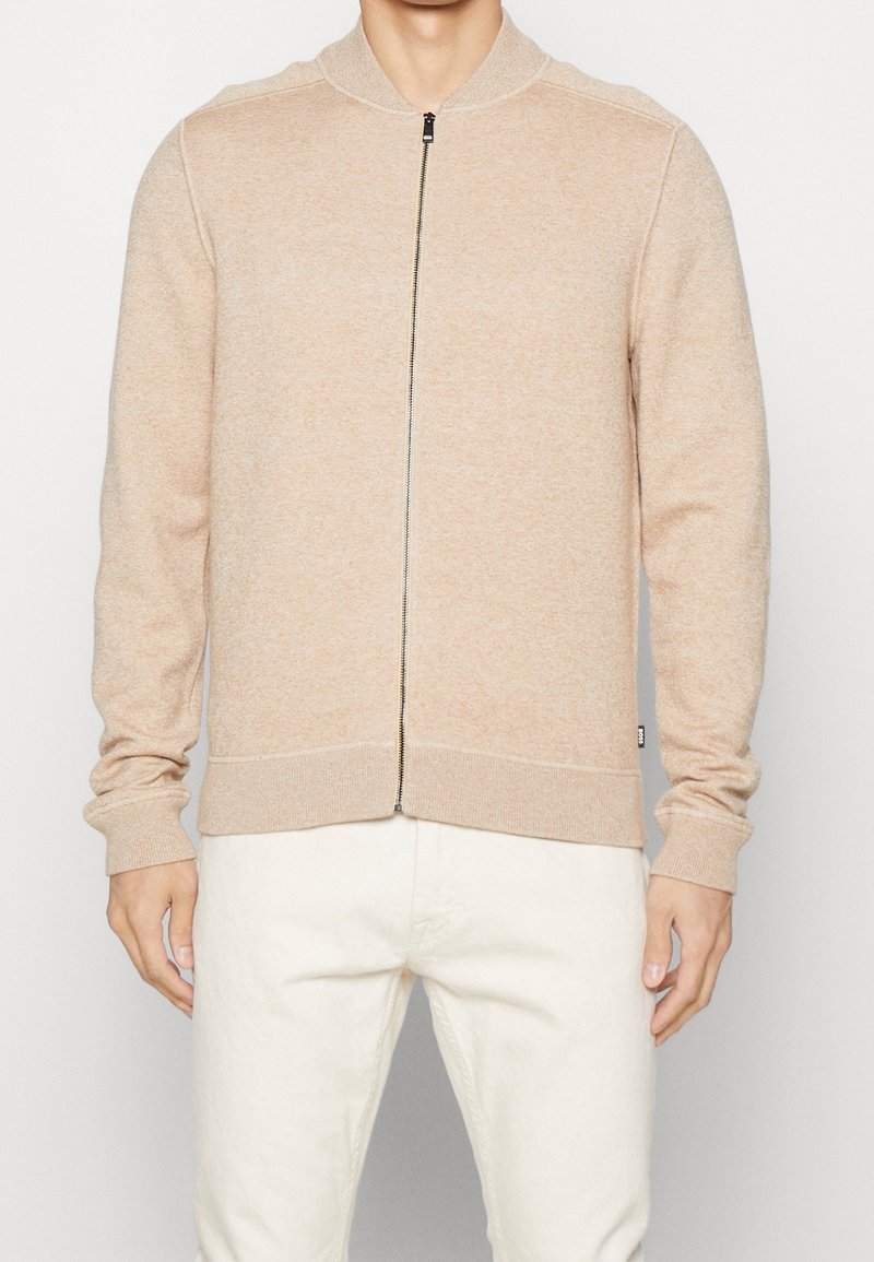 Sudadera beige con cremallera y cuello y puños acanalados, que presenta una tela de punto texturizada y una cremallera de metal visible en el frente.