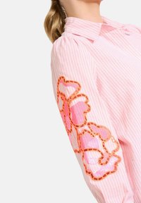 Šviesiai rožinės spalvos dryžuota marškinėliai, su išsiuvinėjimais gėlėmis oranžinės ir rožinės spalvos kairiajame rankove; minkšta medžiaga su apykakle.