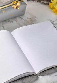 Šedá pouzdro na tužky se zlatými akcenty ve tvaru včely vedle prázdného otevřeného notebooku na mramorovém povrchu. Notebrook má hladké, bílé stránky.
