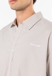 Camicia a righe beige e bianca, con maniche lunghe, colletto e un piccolo logo bianco ricamato sul petto.