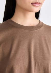 Camiseta de algodón marrón con cuello redondo. La textura es suave y lisa, con un diseño de hombros sin costuras.