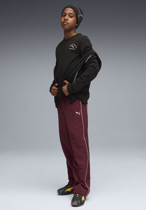 Svart långärmad skjorta med vit logotyp, vinröda byxor med vita sidosträck, och svarta sneakers med gula detaljer, bärs med en svart mössa.