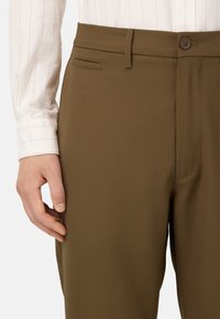 Pantaloni marroni su misura realizzati in tessuto morbido. Presentano una chiusura con un singolo bottone, una tasca laterale e un design frontale piatto.