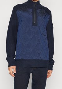 Polo bleu marine avec zip au quart, présentant un devant matelassé et des manches côtelées texturées, fabriqué à partir de matériaux contrastants.