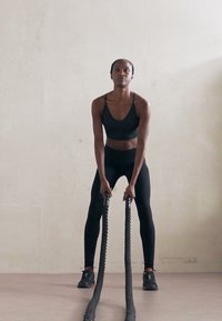 Atleta w czarnym biustonoszu sportowym i legginsach, trzymająca grube czarne liny bojowe, stojąca na gładkiej podłodze z jasną ścianą w tle.