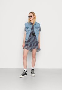 Colete de denim sobre um vestido azul estampado com bainha em renda, combinado com sapatilhas pretas de cano alto e óculos de sol redondos.