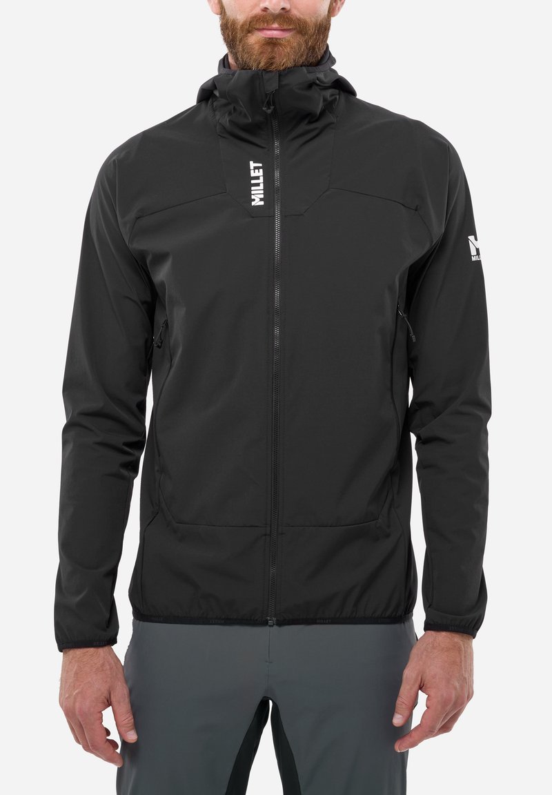 Homme portant une veste zippée noire avec un logo sur la poitrine et la manche, assortie à un pantalon gris foncé, debout devant un fond uni.