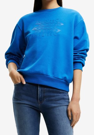 Femme portant un sweat-shirt bleu avec un logo ondulé en relief et un jean bleu, une main dans la poche, sur un fond blanc uni.