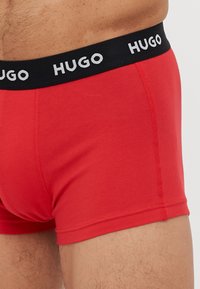 Boxers rouges avec une ceinture noire portant le logo "HUGO" en blanc. Le tissu semble doux avec des détails de couture visibles.